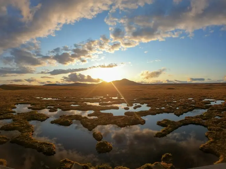 Vibrant sunset over a mountainous landscape, with sky reflected in patchy water. Clouds have a golden hue, creating a serene and expansive scene.
